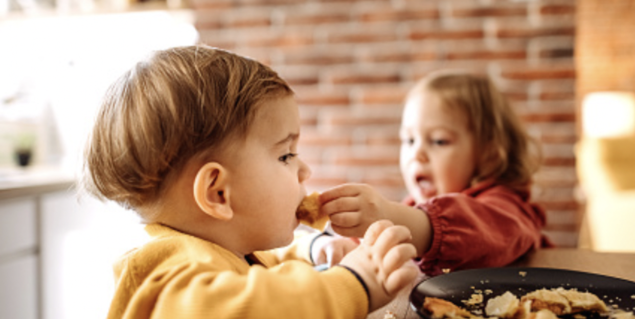 a young child leans across the table and shares some food with another young child. I wonder if it was nice food, I hope so. It's a sign of being as one